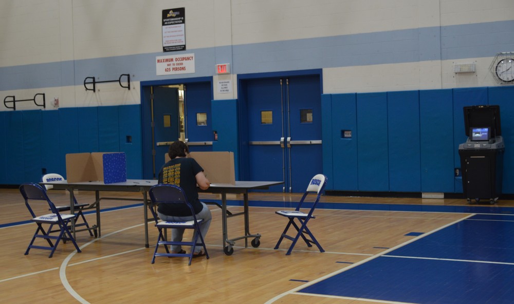 A person sits at a table filling out a ballot with the ballot reader at right