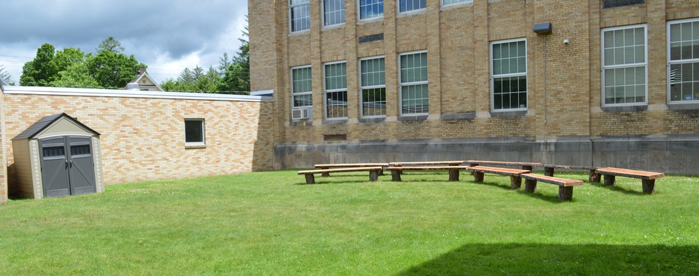 Two rows of benches on a curve sit in the outdoor classroom with a storage shed to the left