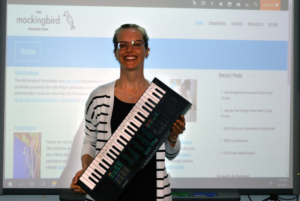 A woman holds an old electric piano in front of  a screen with the Mockingbird Foundation's home page displayed